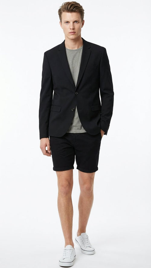 Men’s black summer business suit features a fitted black blazer with two buttons and notched lapels, paired with matching black shorts. The blazer has two front pockets and structured shoulders, while the shorts are knee-length with a cuffed hem. A gray t-shirt is worn underneath the blazer.