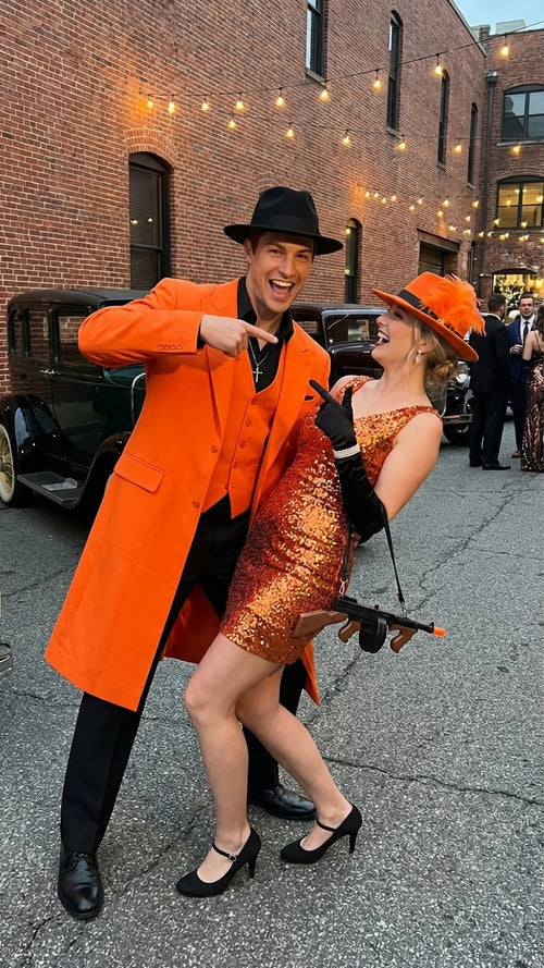 Orange zoot suit with black shirt and pants. Elongated, smooth fabric suit with a sharp collar and lapels. Buttoned black shirt and straight-cut pants complete the look.