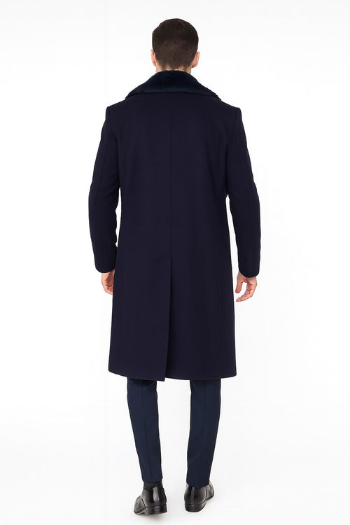 Navy blue men's overcoat with faux fur collar. Long, structured winter coat extending past the waist. Features tailored sleeves, straight back, and smooth, textured material. Paired with navy trousers and black shoes.
