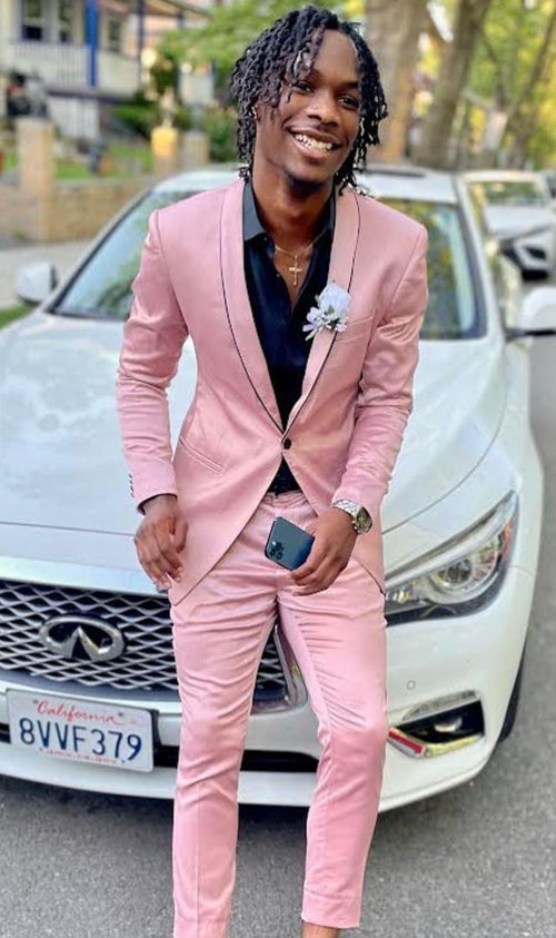 Men's slim fit pink tuxedo with black trim. Tailored jacket, notched lapel, two pockets, and matching trousers. Smooth, slightly shiny fabric. Black trim on lapel and pockets. Paired with a black shirt.