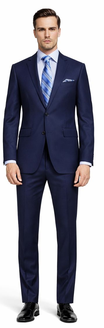 Navy blue, two-button suit (slim/modern fit). Structured jacket, straight-cut trousers. Smooth, refined fabric. Jacket has two front pockets. Paired with light blue shirt, striped tie.
