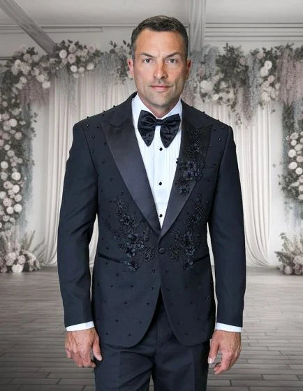 The Statement mens 2 piece modern fit tuxedo features a black fabric with floral appliqué detailing. The jacket has a structured silhouette with peak lapels and is adorned with embroidered elements across the front. The ensemble includes matching trousers and a white dress shirt.