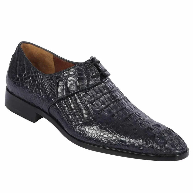 Navy crocodile calfskin slip-on shoes with pointed toe and low profile. Textured surface, dark navy leather. Simple strap with buckle. Black sole, slight heel. Sleek design.