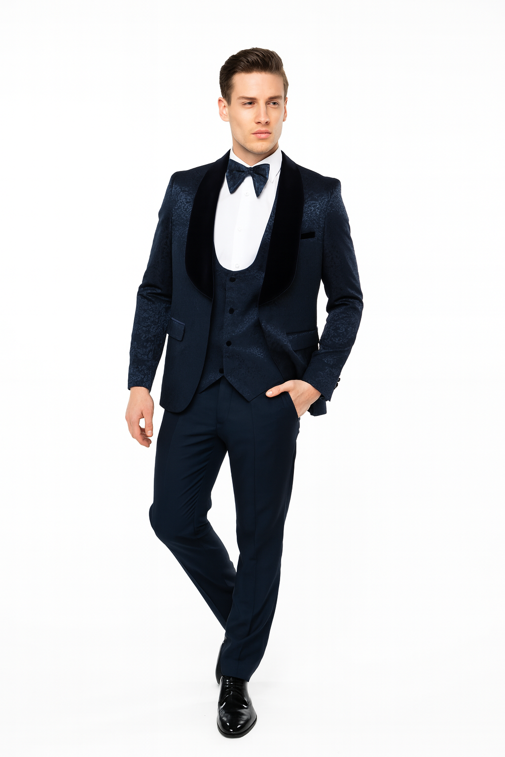 Navy jacquard tuxedo with black velvet shawl lapel. Tailored jacket, structured shoulders, fitted waist. Flat-front trousers. White shirt and bow tie complete the formal look.
