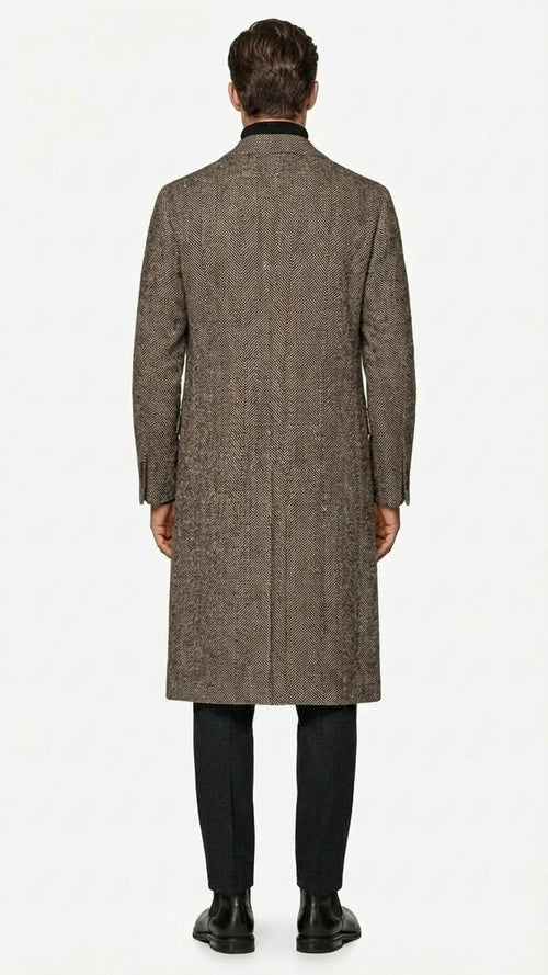 Men's black & grey herringbone tweed overcoat. Long, straight, single-breasted wool blend. Notched collar, side vents, & visible stitching. Structured, textured design.
