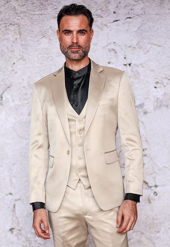 Men's champagne slim-fit suit in shiny satin. Three-piece set: jacket (single-button, notched lapel), vest, and trousers. Jacket has side pockets. Worn with a contrasting black shirt.
