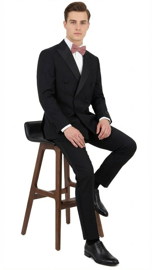 Men's classic black double-breasted tuxedo in 100% Italian wool. Features peak lapels, tailored fit, structured shoulders. Paired with black shoes and a light pink bow tie for contrast.