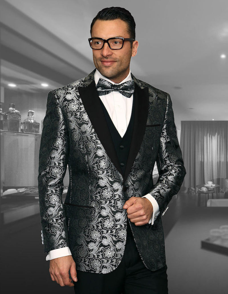 Black one-button peak lapel tuxedo with silver floral pattern and sheen. Includes vest and bow tie. Features a structured silhouette and ornate design. Non-functional sleeve buttons.
