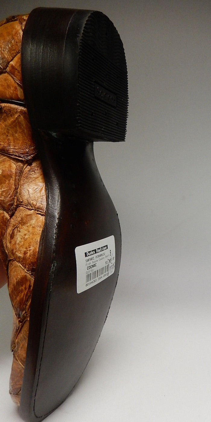 Cognac-colored safari cowboy boots crafted from pirarucu fish scales. Black, rubberized sole with a subtle heel. Features smooth leather side panels, stitching, and product label.
