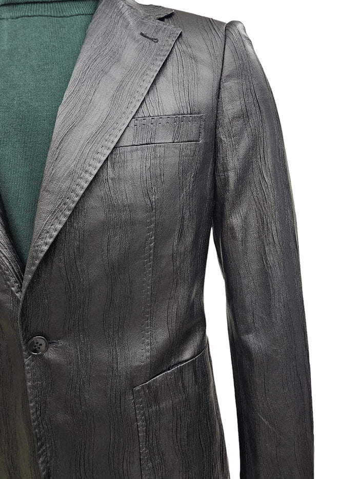 Black men's slim fit suit. One-button closure, notch lapels, textured finish with wave pattern. Jacket has chest/lower pockets. Worn with green garment.
