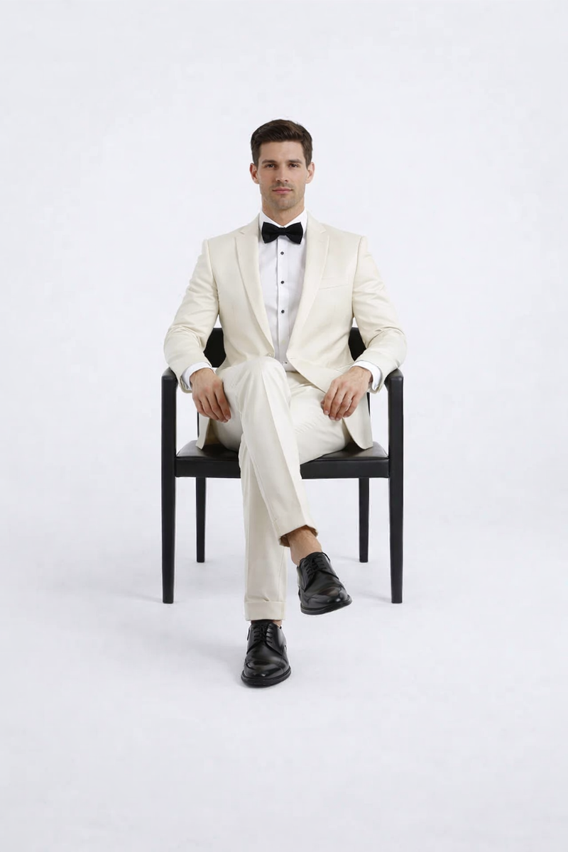 Ivory peak lapel slim fit tuxedo: single-breasted jacket, lightweight fabric, smooth finish. Includes matching trousers, white shirt, and black bow tie. Structured silhouette.
