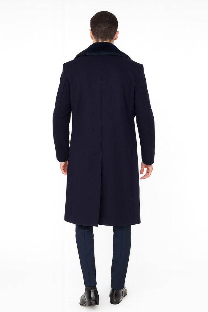 Navy blue men's overcoat with faux fur collar. Long, structured winter coat extending past the waist. Features tailored sleeves, straight back, and smooth, textured material. Paired with navy trousers and black shoes.
