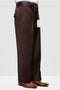 Italian wool wide-leg dress pants in brown. Straight, loose fit with front pleats. Smooth, structured fabric. Tailored cuffs rest above dress shoes, emphasizing the wide silhouette. Features side pockets and a waistband tag.

