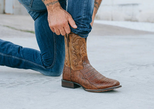 Dan Post Men's Kingly boots: Genuine caiman belly leather with a square toe and tall shaft. Brown upper with subtle pattern and decorative stitching. Stacked heel, textured sole for a sleek, structured look.
