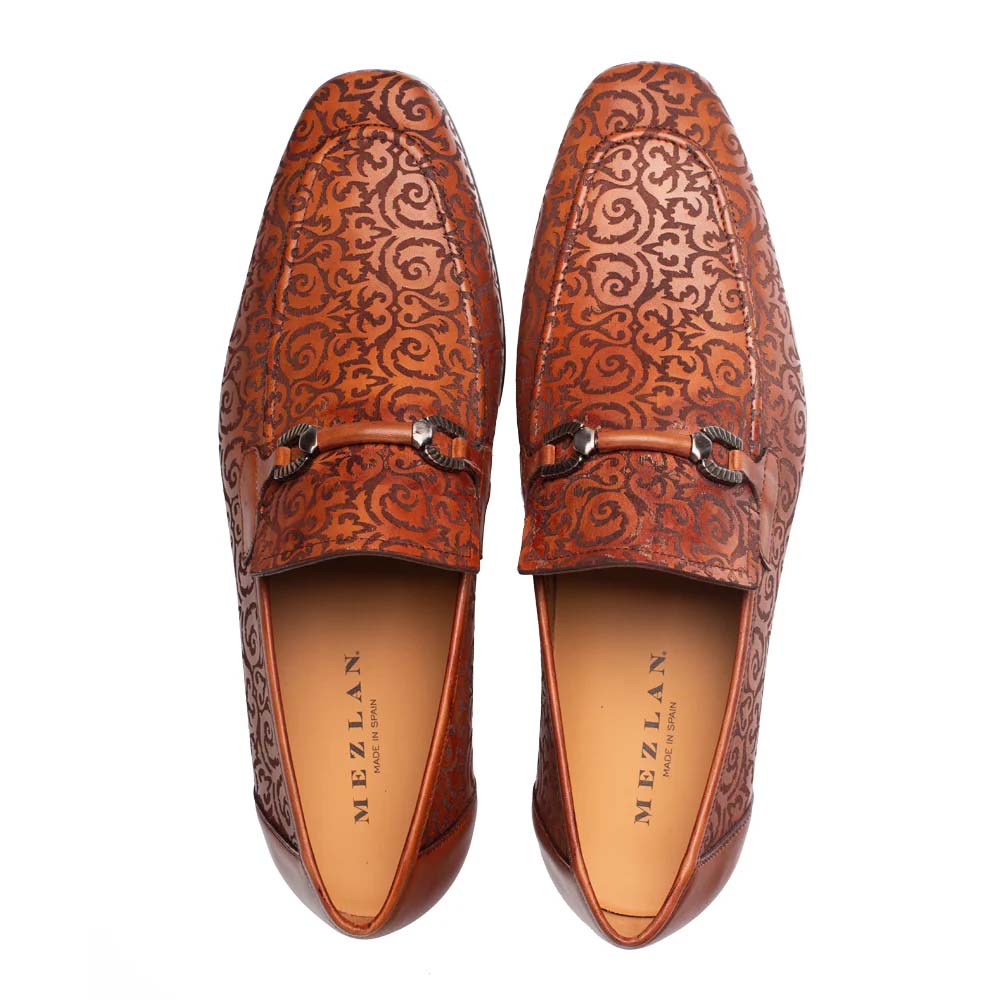 Cognac Mezlan Alcaraz slip-ons. Leather upper with laser-cut swirling pattern. Elongated shape, rounded toe, and contrasting interior. Decorative front elements add detail. Smooth, flat design.
