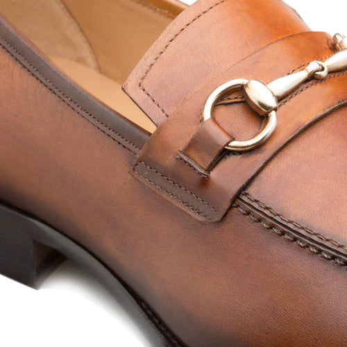 Cognac Mezlan loafers: smooth leather, rounded toe, decorative strap with metal buckle. Sturdy black rubber sole, visible stitching. Warm medium-brown color.
