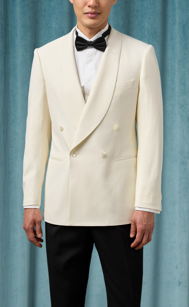Ivory double-breasted men's tuxedo jacket. Fitted silhouette, shawl lapel, and two rows of buttons. Smooth, lightweight ivory fabric with structured shoulders, front pockets, and buttoned cuffs. Tailored look.
