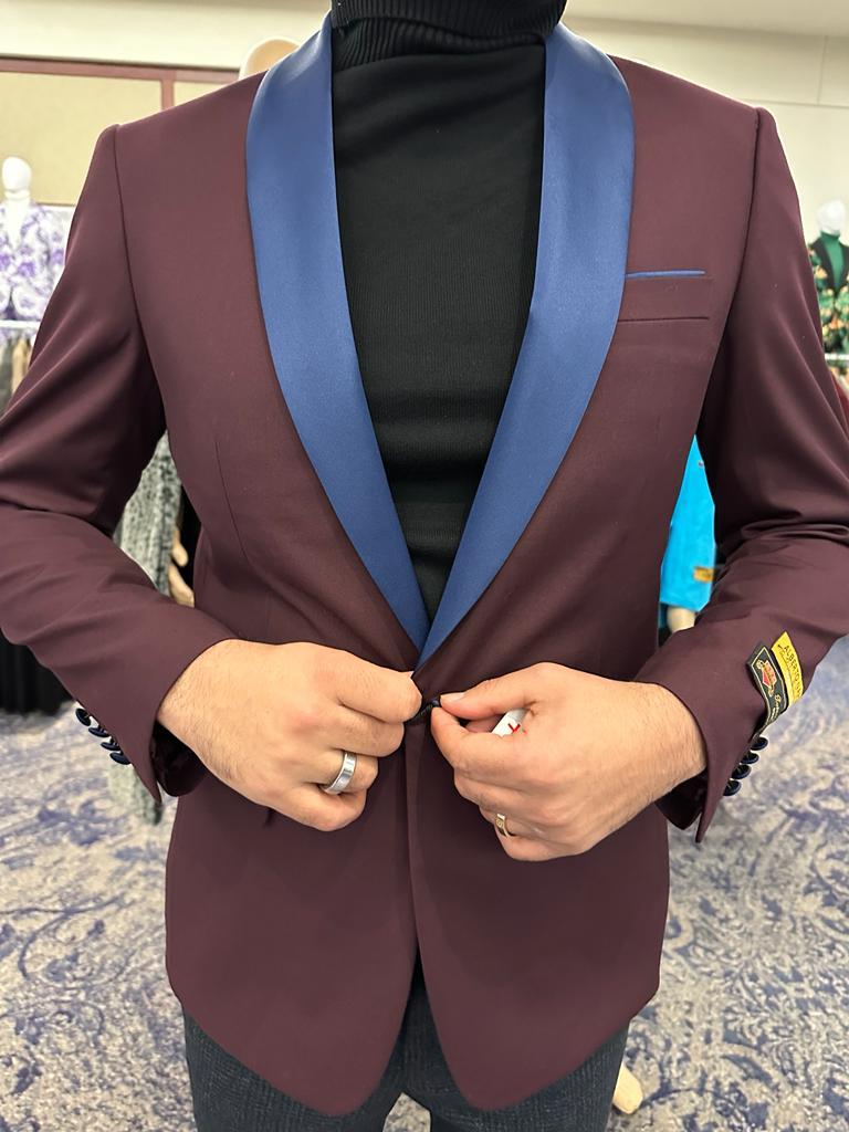 Men's burgundy tuxedo blazer with blue satin shawl collar. Fitted silhouette, single-button closure, chest pocket, and long sleeves. Smooth, likely sheen fabric.
