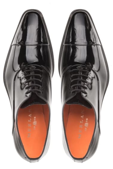 Black patent leather Oxfords with pointed toe, lace-up design, and orange lining. Featuring a reflective surface, slim textured sole, and rounded heel for a polished look.
