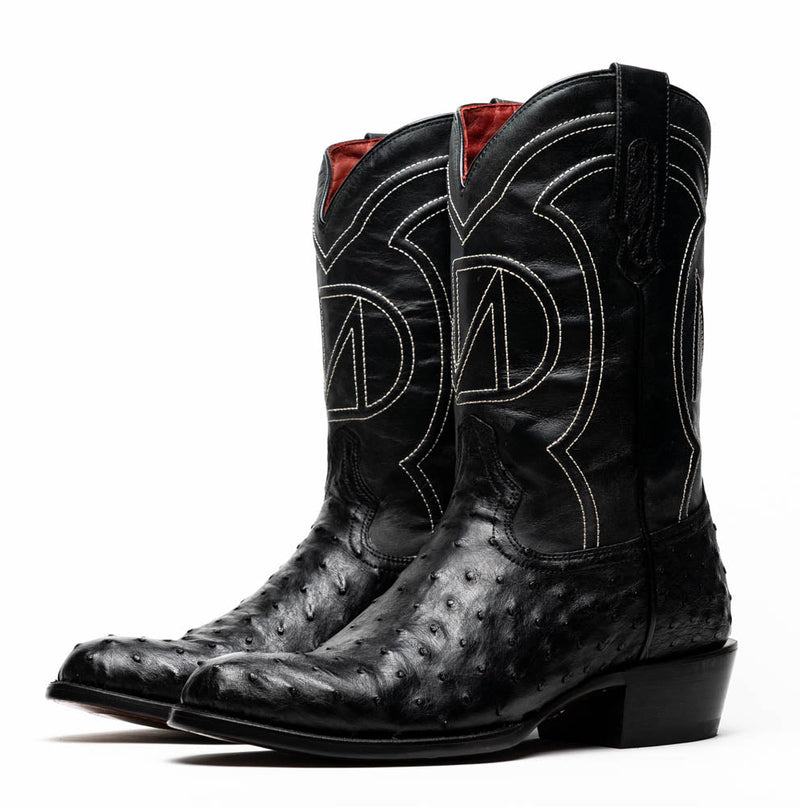 Black ostrich quill cowboy boots by Marco Di Milano. Tall shaft with curved top, textured leather, round toe. Moderate stacked heel. White stitching design, red lining, pull tabs. Ostrich skin texture.
